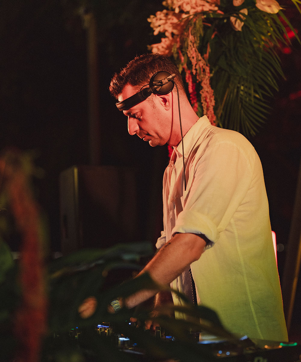 A DJ wearing headphones and a light-colored shirt stands at a mixing console, focused on his equipment. He is surrounded by lush greenery and flowers, with warm lighting creating a vibrant atmosphere.