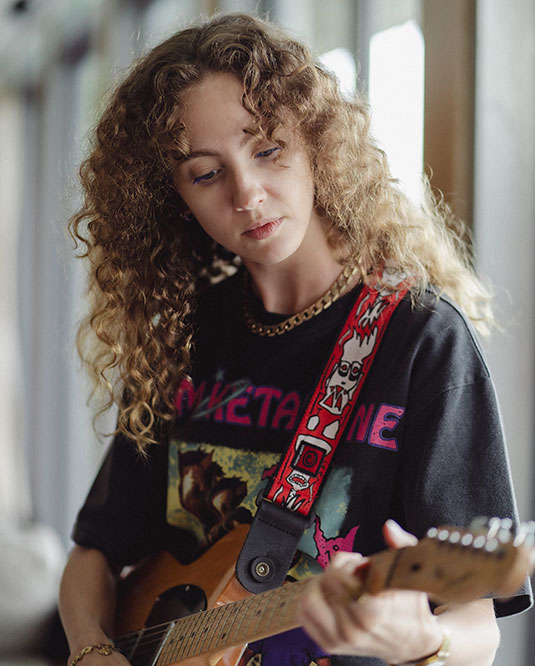 A young woman with curly hair plays an electric guitar indoors. She wears a black graphic T-shirt and a patterned red guitar strap, looking down at the instrument with a focused expression.
