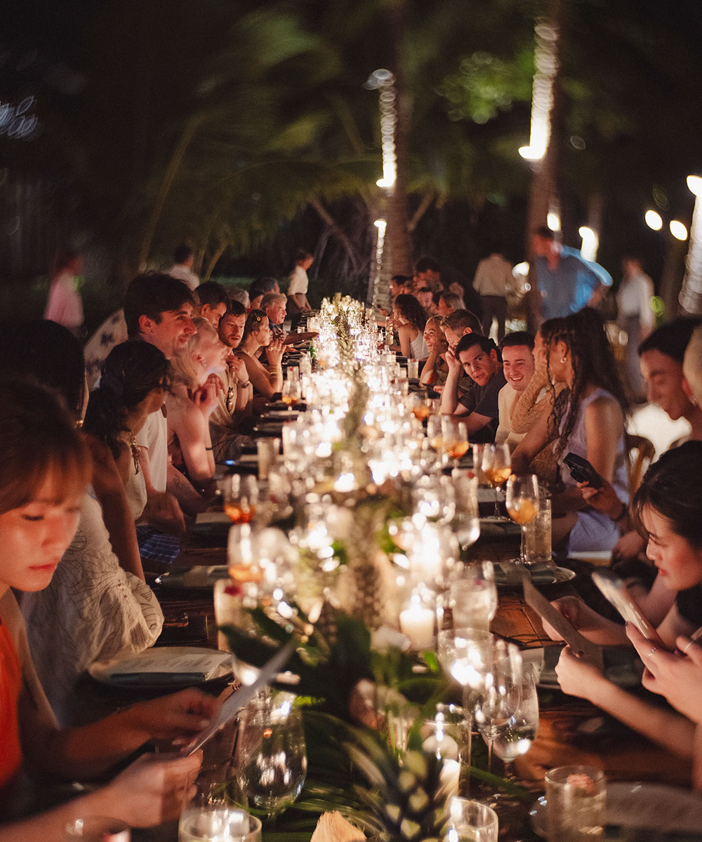 A long outdoor dinner table is filled with people enjoying a meal together at night, surrounded by greenery and illuminated by candles and string lights, creating a warm and festive atmosphere.