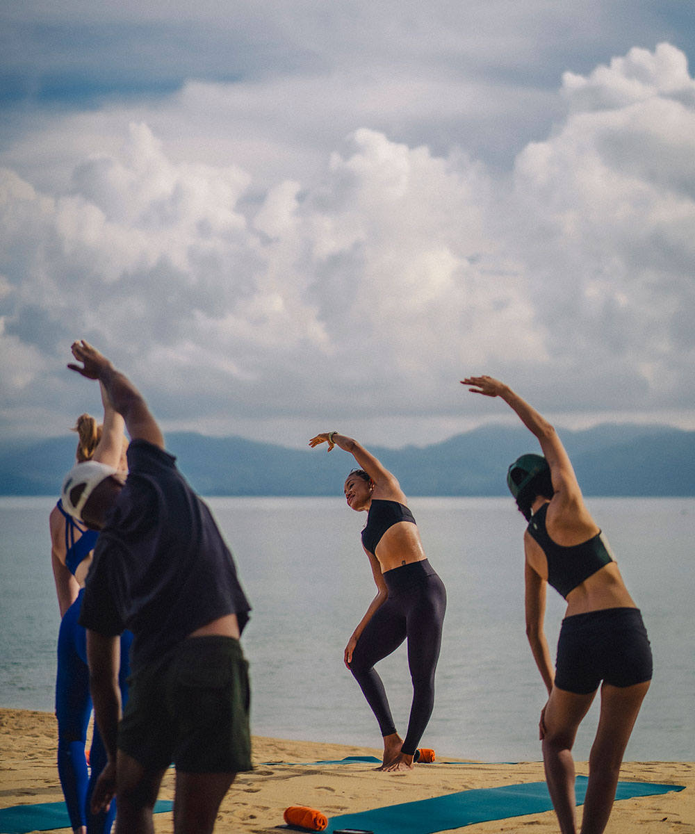 A group of people practice yoga on mats on a sandy beach, stretching with one arm overhead. The sea and mountains are visible in the background under a cloudy sky.