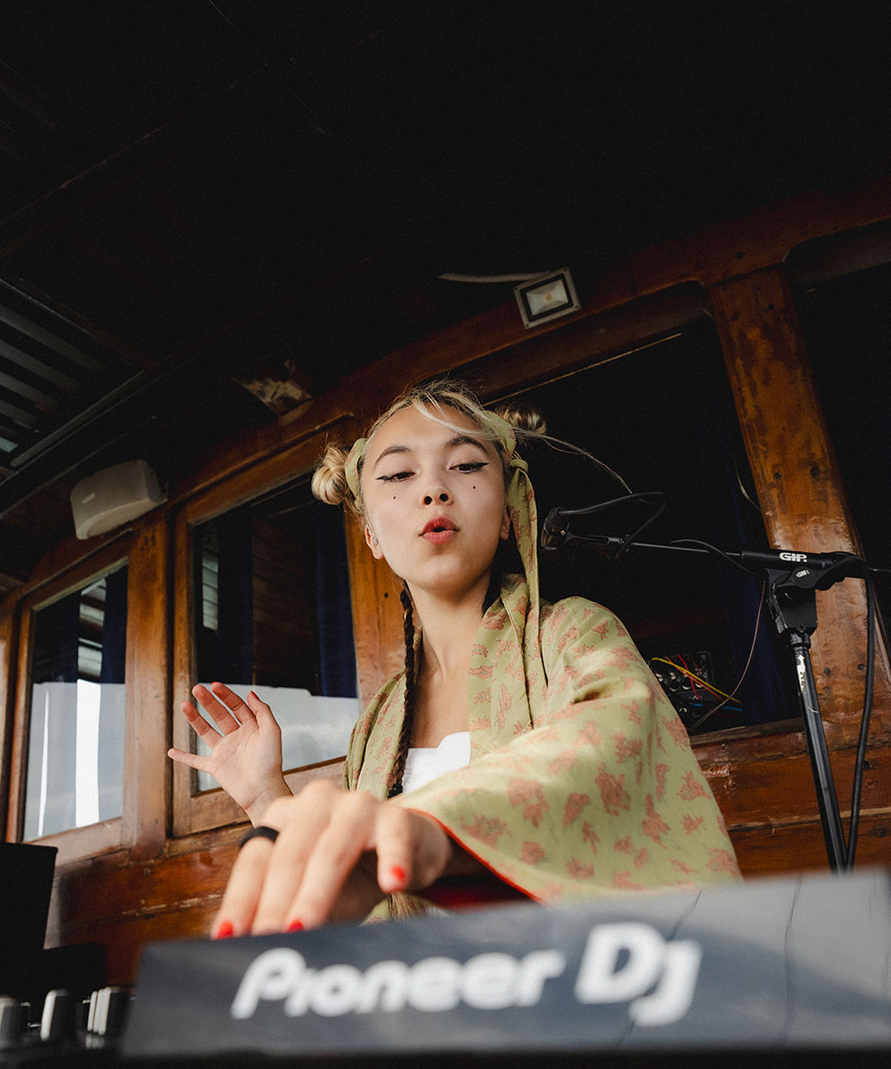 A young woman with space buns and face makeup is DJing on a Pioneer DJ controller. She wears a green patterned kimono and gestures with one hand, with wooden paneling and a microphone stand in the background.