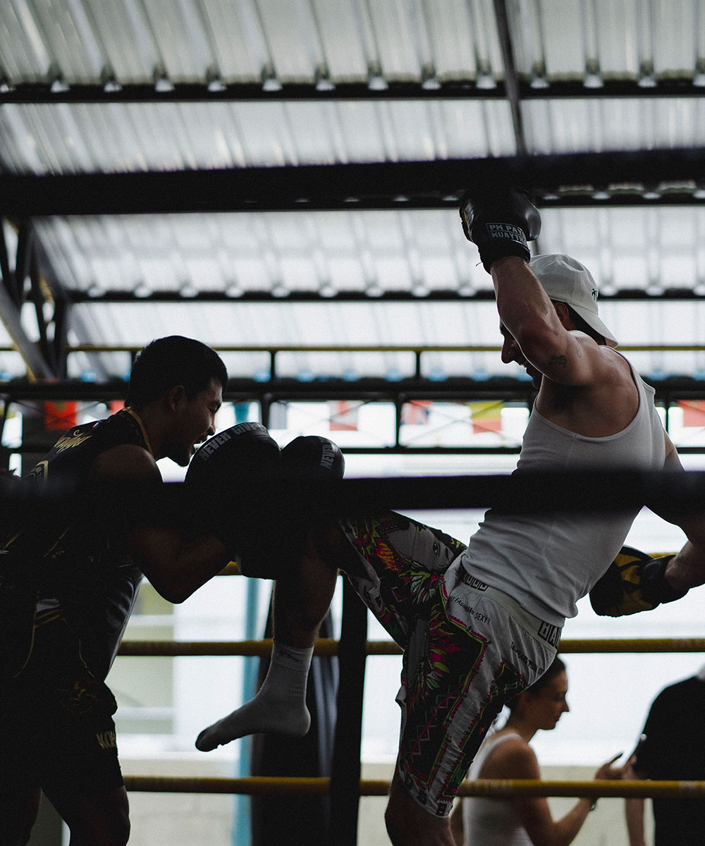 Two people spar in a boxing ring; one throws a knee while wearing patterned shorts and a white tank top, the other blocks with padded gloves. The background shows a woman standing outside the ring.
