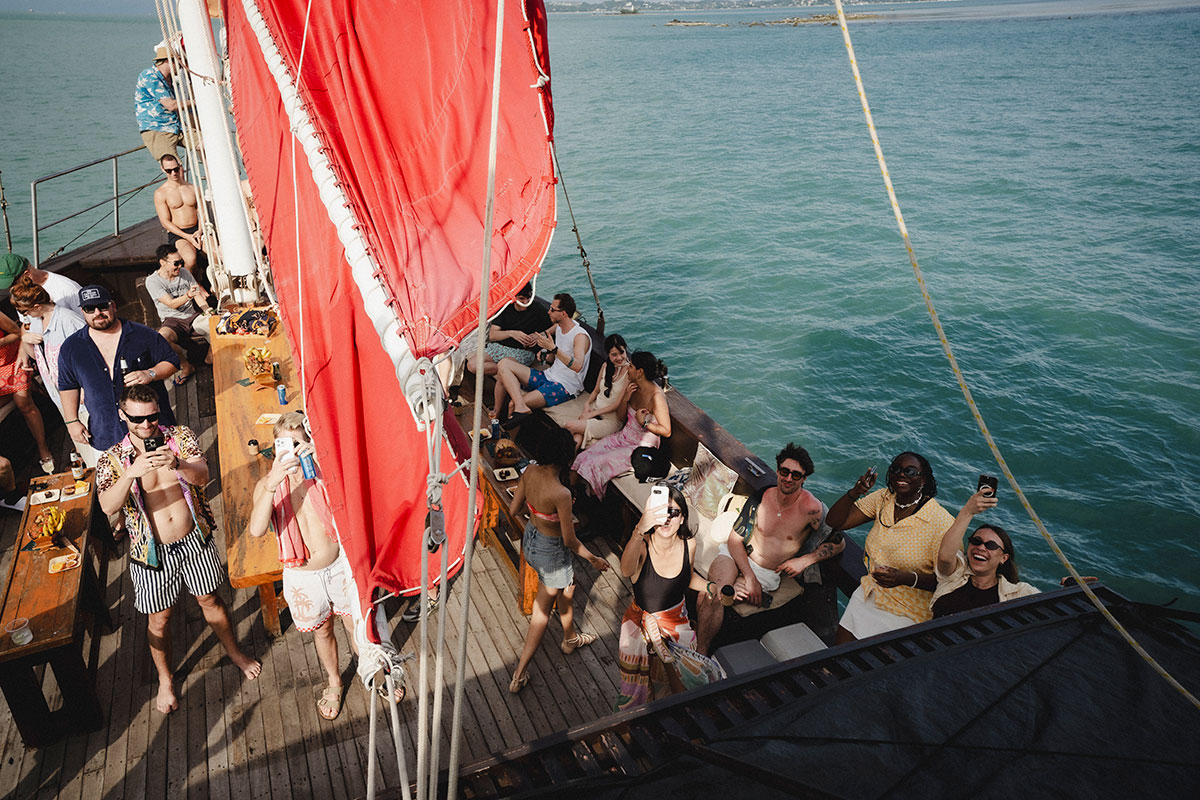 A group of people in swimwear relax and enjoy drinks on a wooden sailboat with a large red sail, floating on clear blue water under the sun. Some people are smiling and raising glasses toward the camera.