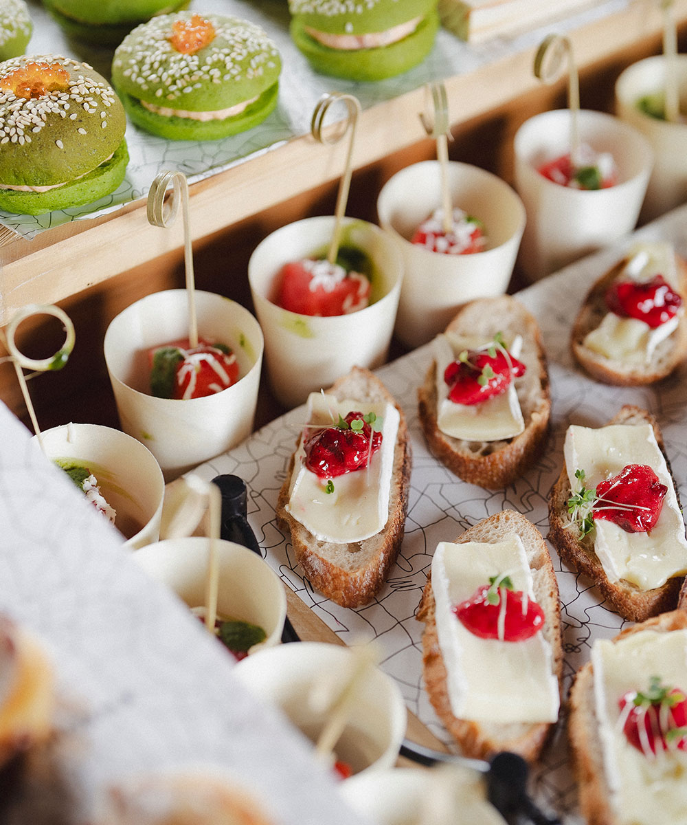An assortment of elegant appetizers, including green macarons, vegetable cups, and slices of bread topped with cheese, herbs, and red garnish, arranged on a table for a catered event.