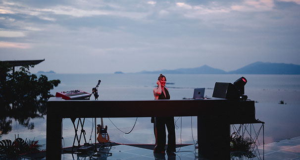 A DJ stands behind a large outdoor setup with electronic equipment, performing near a body of water at sunset, with mountains visible in the distance under a cloudy sky.
