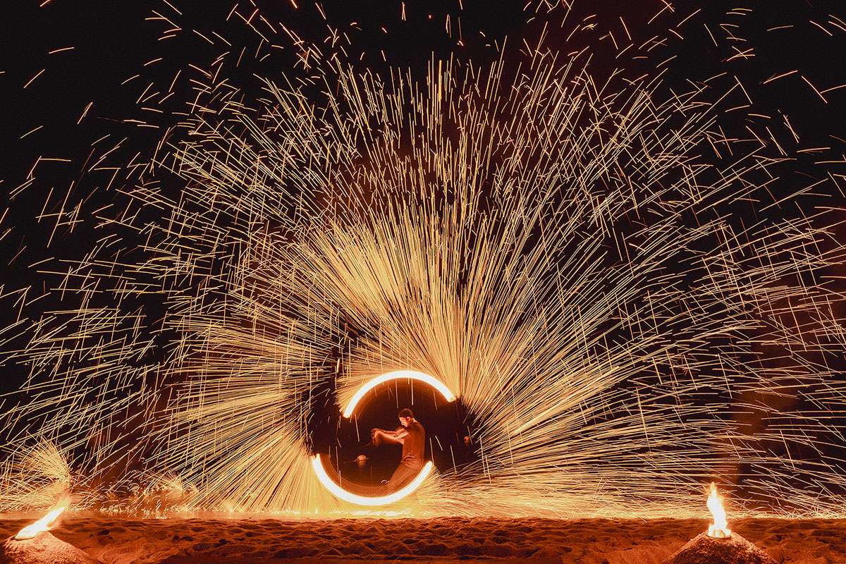 A performer spins a fire staff, creating a circle of light and a burst of golden sparks against a dark background, with two small fires burning on the ground in front.