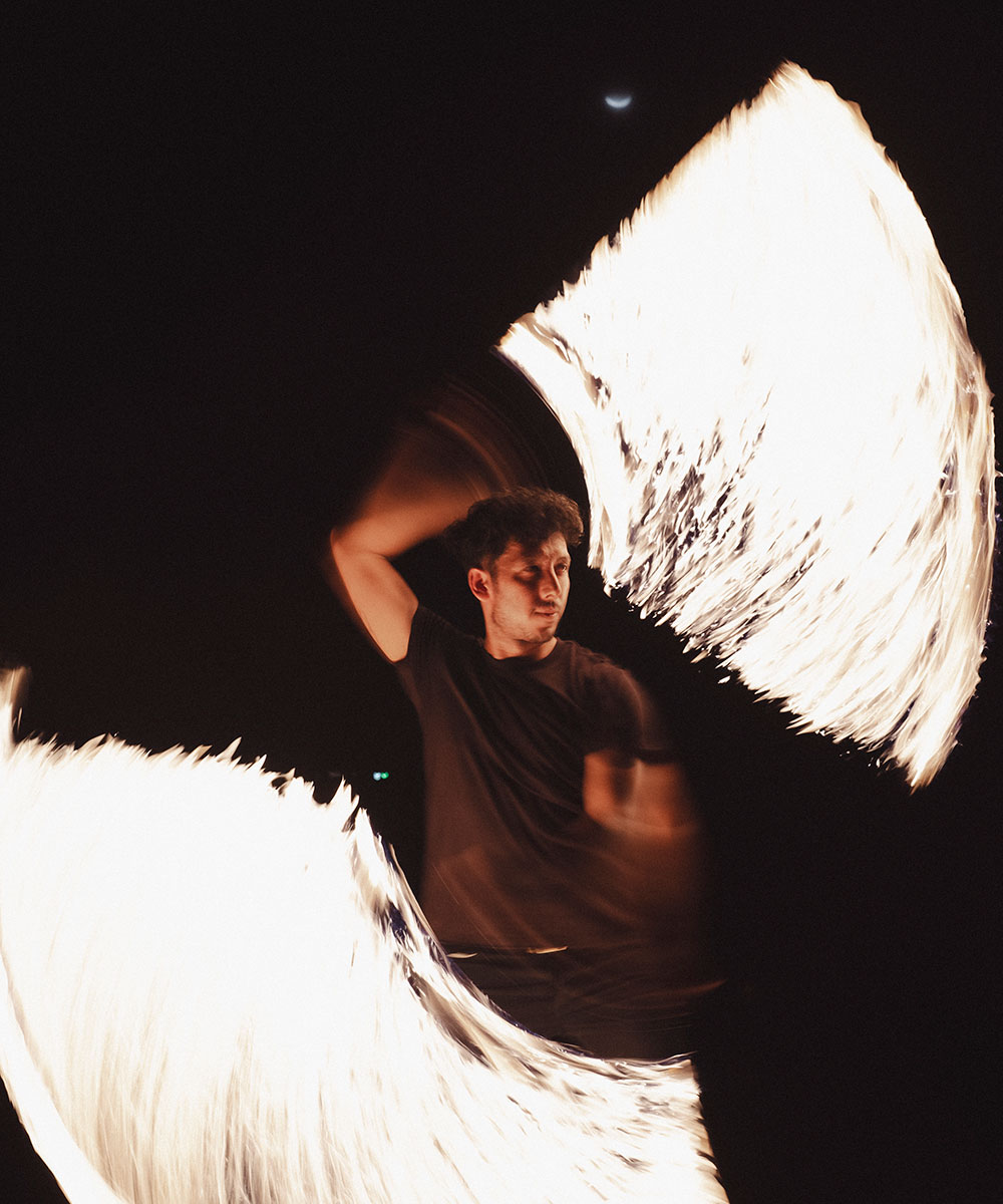 A person performs a fire dance at night, skillfully spinning flaming props that create bright, swirling trails of light against a dark background.