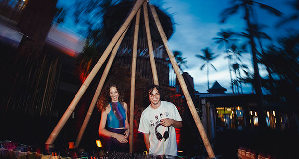 Two DJs stand behind a mixing console at an outdoor event during dusk, surrounded by palm trees and dim, colorful lighting, with a wooden teepee structure framing them.