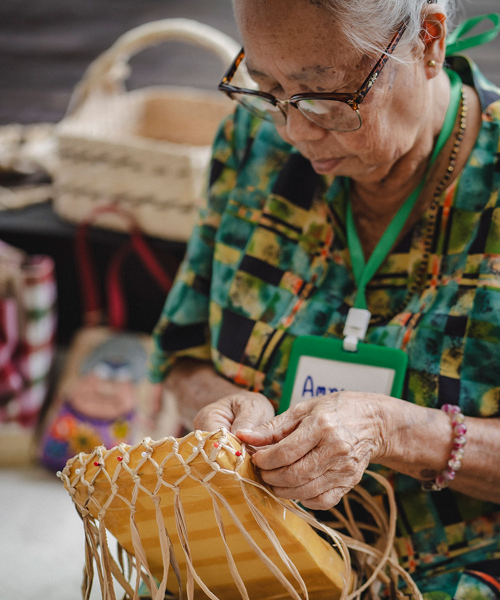 An elderly woman wearing glasses and a green patterned shirt weaves a basket from straw. She is focused on her work, surrounded by other woven baskets and crafts in the background.