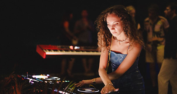 A young woman with curly hair wearing headphones and a blue dress DJs at a brightly lit console, with a keyboard and blurred people in the background.