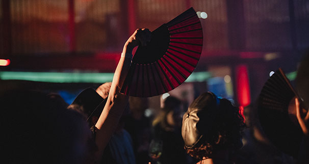 A person holds up an open black and red hand fan in a dimly lit indoor crowd, with warm red and yellow lights in the background.