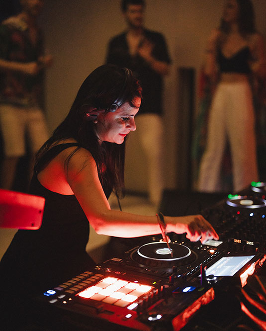 A female DJ in a dark setting is mixing music on a DJ controller, illuminated by red and orange lights. People are standing and dancing in the blurred background.