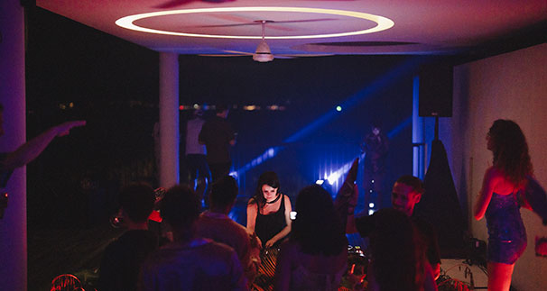 A group of people dance in a dimly lit room with colorful lights and a DJ at the center. The atmosphere is lively, with partygoers enjoying music under a modern ceiling fixture.