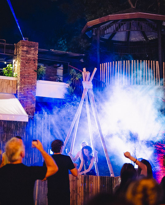 A group of people at an outdoor night event watch a woman near a wooden structure surrounded by blue and white stage lights and smoke effects, creating a lively and festive atmosphere.