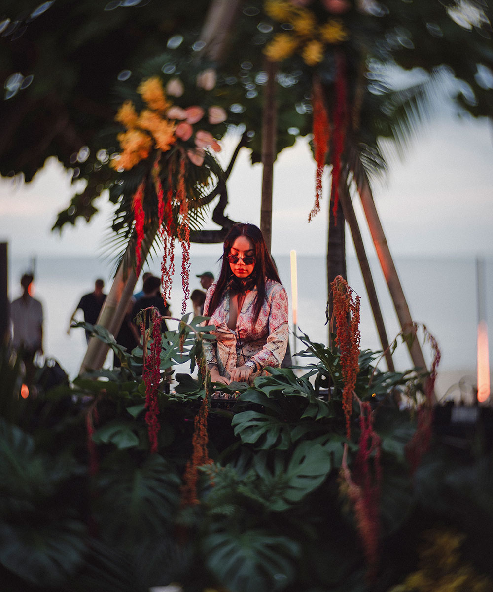 A woman wearing sunglasses DJs outdoors behind lush green leaves and hanging red flowers, with the ocean and silhouettes of people visible in the background.