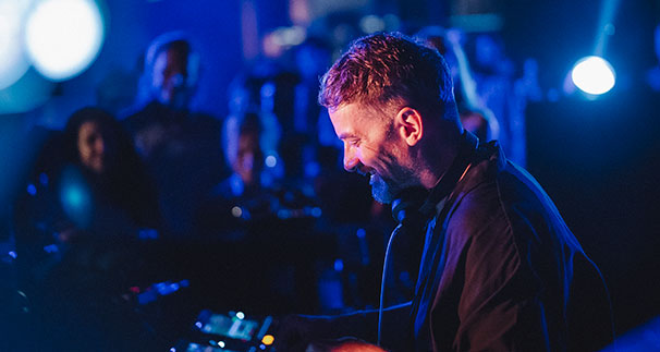 A DJ with short hair and headphones around his neck smiles while performing at a dimly lit club, surrounded by blue lighting and an audience in the background.