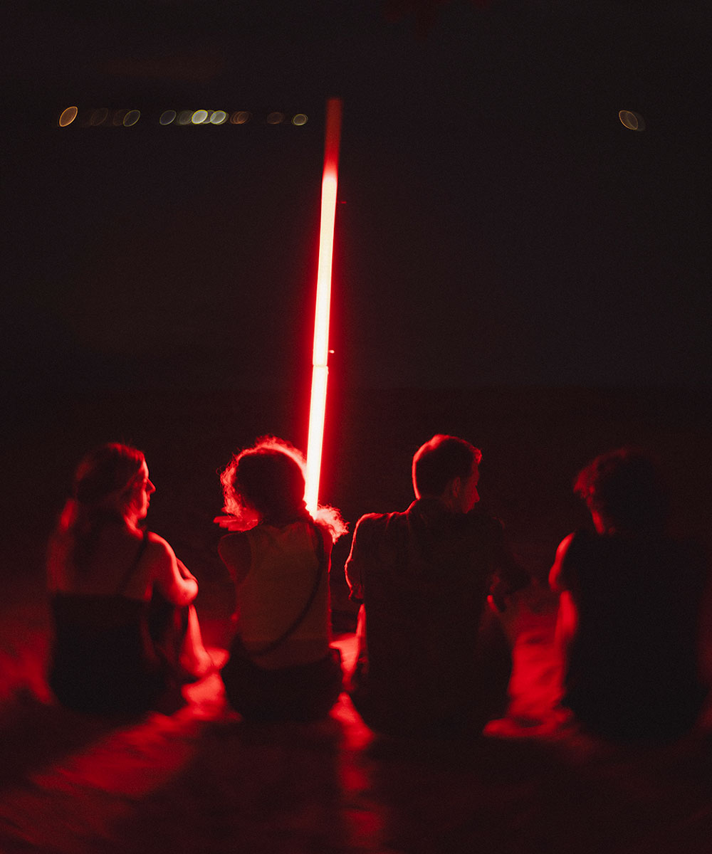 Four people sit on the ground in the dark, illuminated by a vertical red light between them. The scene is dim, and their faces are not clearly visible. Small lights appear in the distant background.