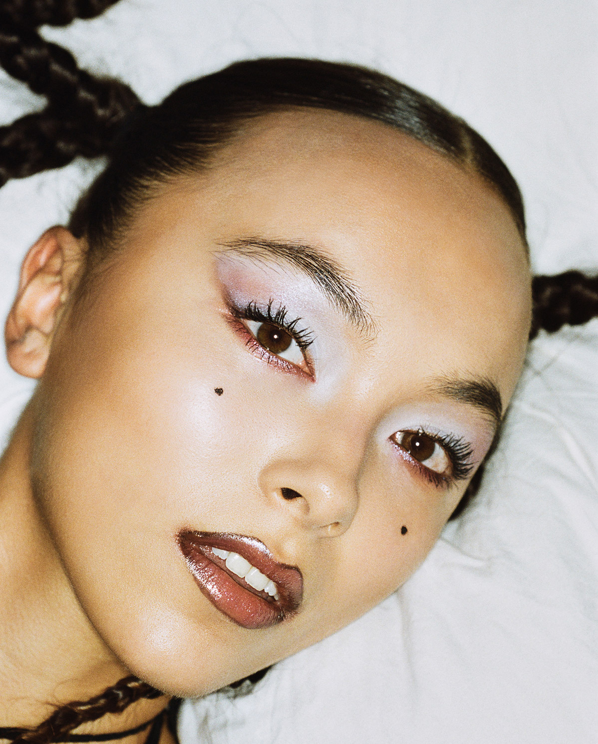 A close-up of a person with glossy lips, shimmery pastel eyeshadow, long eyelashes, and two small black beauty marks on their cheek, lying on a white pillow with hair styled in braids.