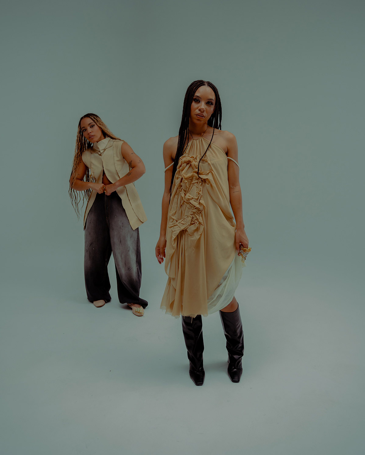 Two women pose in a minimalist studio. One wears a flowing tan dress and black boots, standing in the foreground. The other, in a sleeveless beige top and wide dark pants, stands behind her. The background is plain and light-colored.