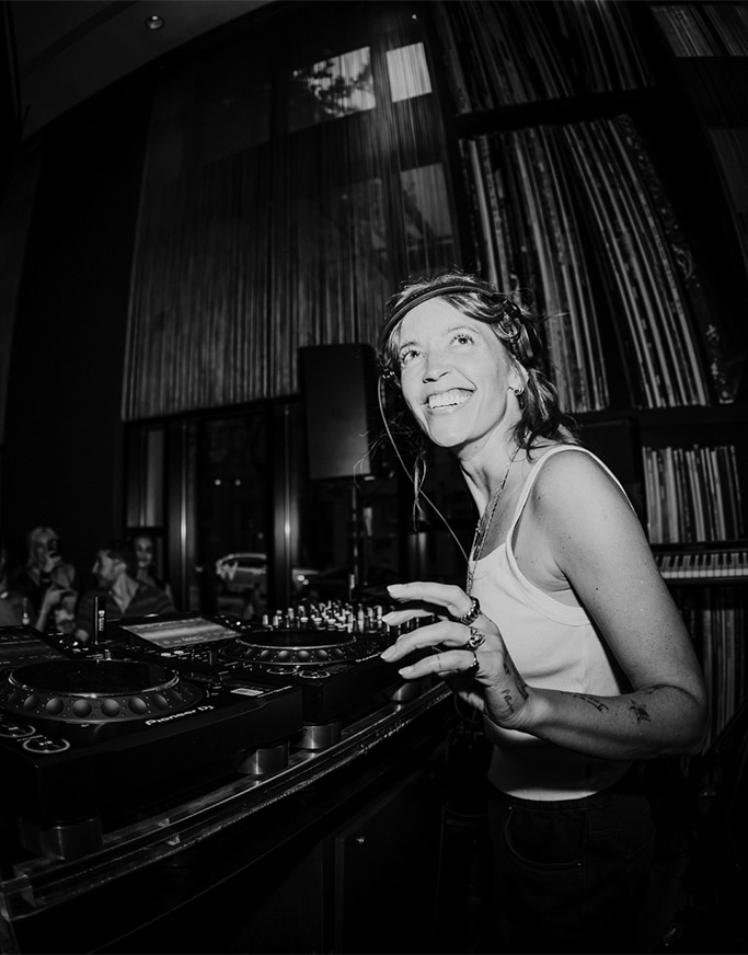 A smiling DJ wearing a tank top and headphones stands behind a mixing console in a dimly lit room, surrounded by tall shelves filled with vinyl records. The image is in black and white.