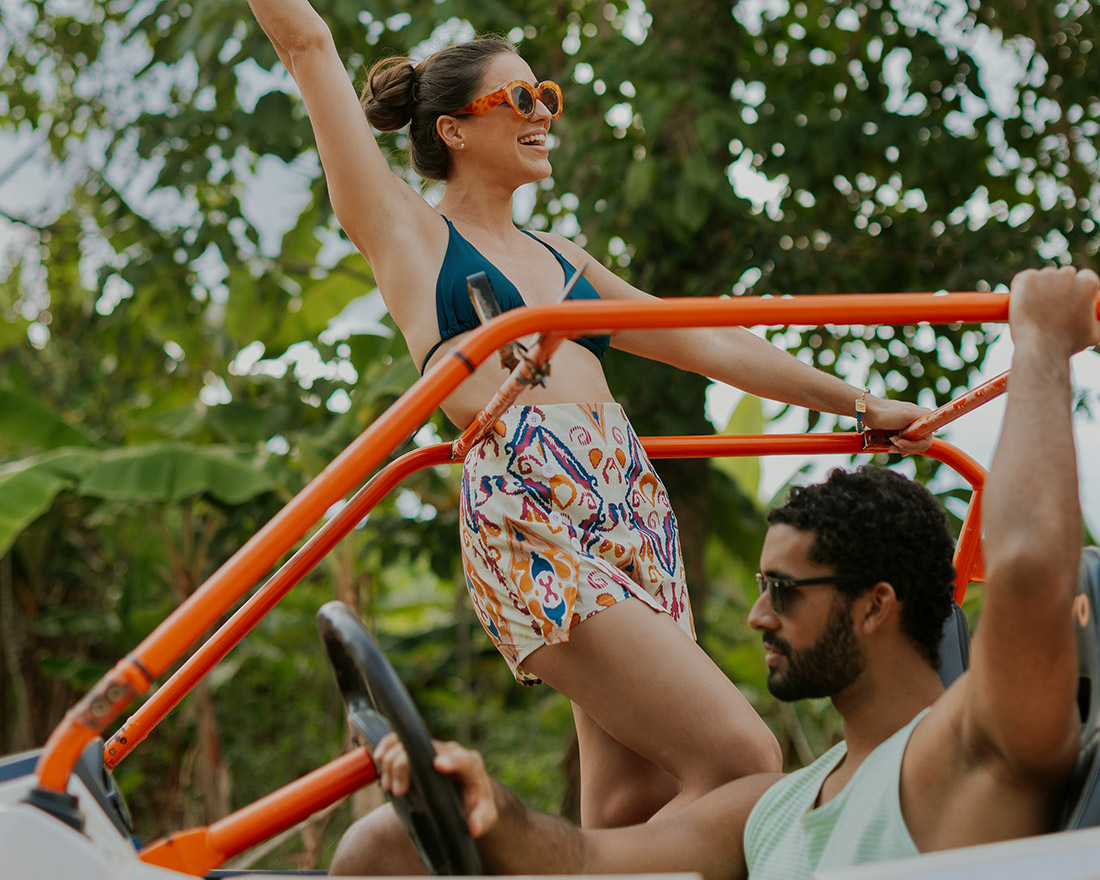 A woman in sunglasses stands in a colorful open-top vehicle, smiling with one arm raised, while a man in sunglasses drives. Lush green plants and trees are in the background, suggesting a tropical setting.