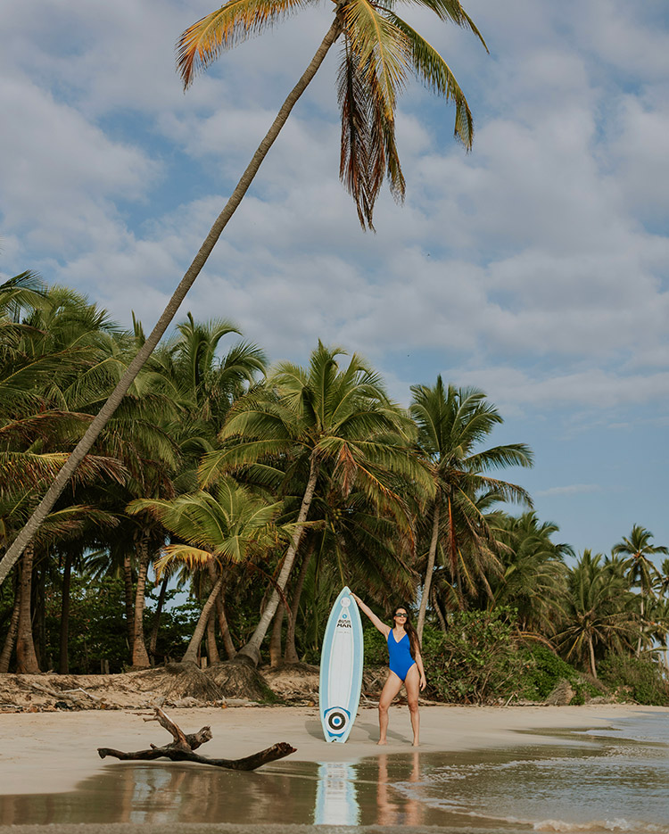 Eine Frau in einem blauen Badeanzug steht an einem Sandstrand neben einem Surfbrett, umgeben von hohen Palmen unter einem teilweise bewölkten Himmel.