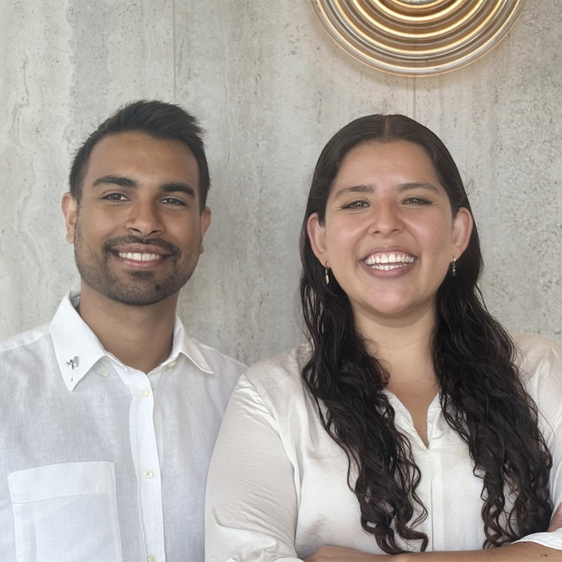 Two people stand side by side, smiling, in front of a textured light-colored wall with a circular decorative light above them. Both wear white shirts and have dark hair.