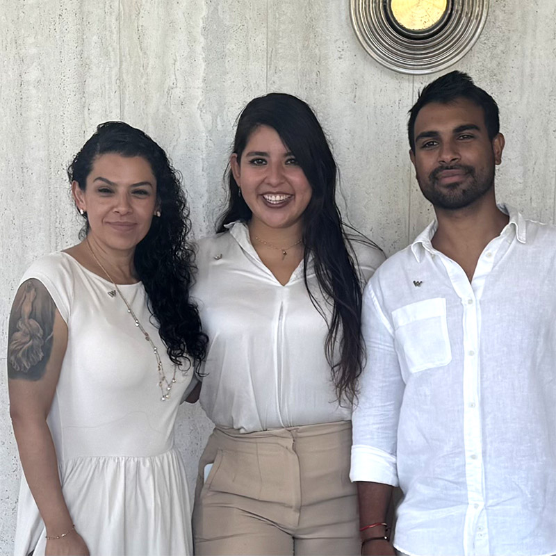 Three people stand side by side, smiling at the camera. They are dressed in light-colored, semi-formal clothing and are standing in front of a light-colored textured wall with a circular wall decor above them.