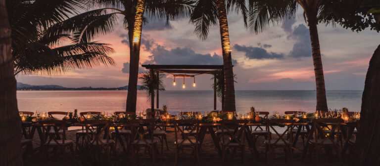 A long, candlelit dining table is set on the beach beneath palm trees at sunset, with a calm sea and colorful sky in the background. Hanging lights add a warm glow to the elegant outdoor setting.