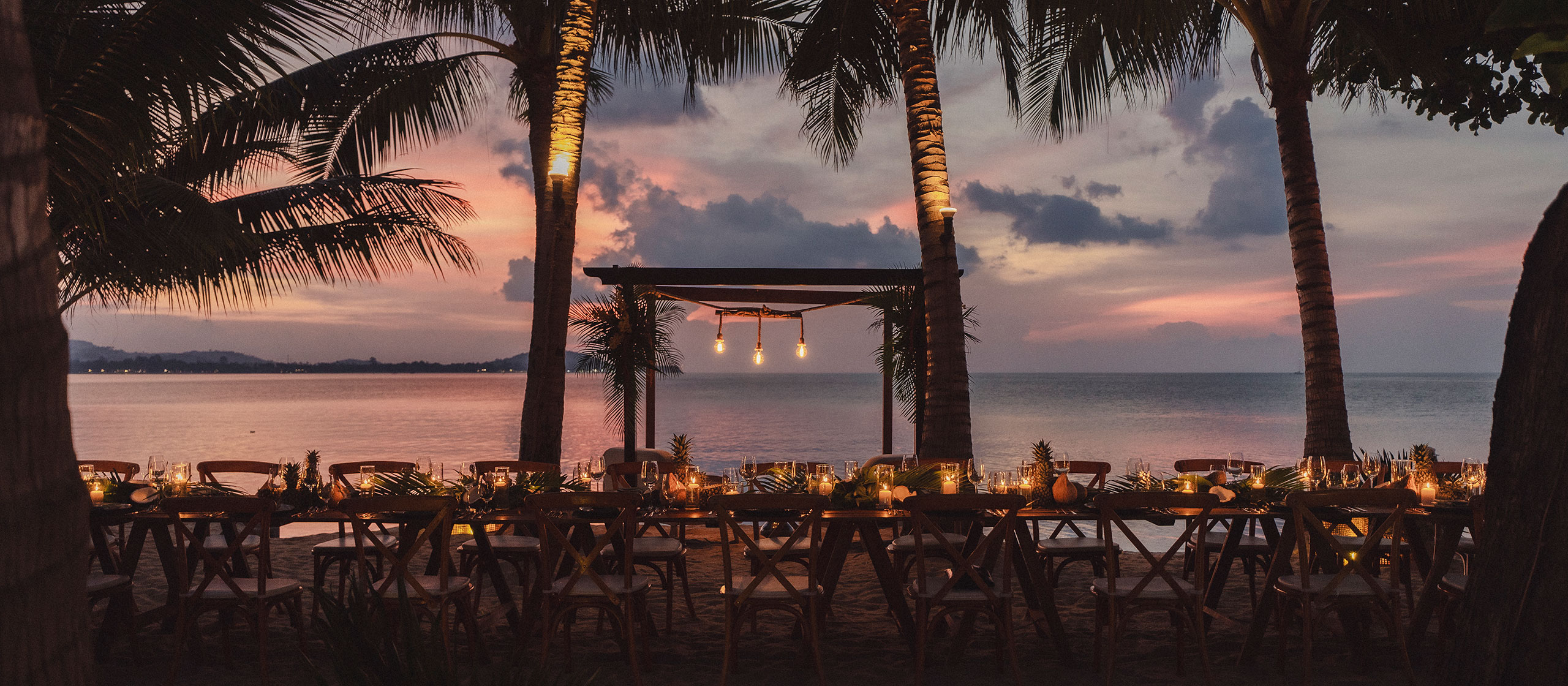 A long, candlelit dining table is set on the beach beneath palm trees at sunset, with a calm sea and colorful sky in the background. Hanging lights add a warm glow to the elegant outdoor setting.