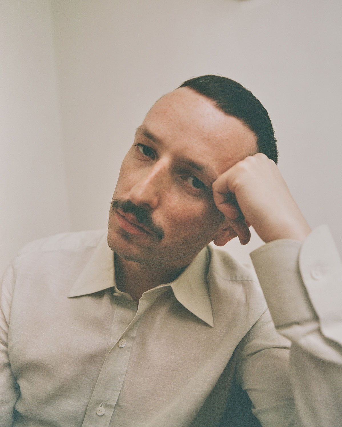 A man with short dark hair and a mustache, wearing a light-colored button-up shirt, rests his head on his hand and looks thoughtfully at the camera against a plain background.