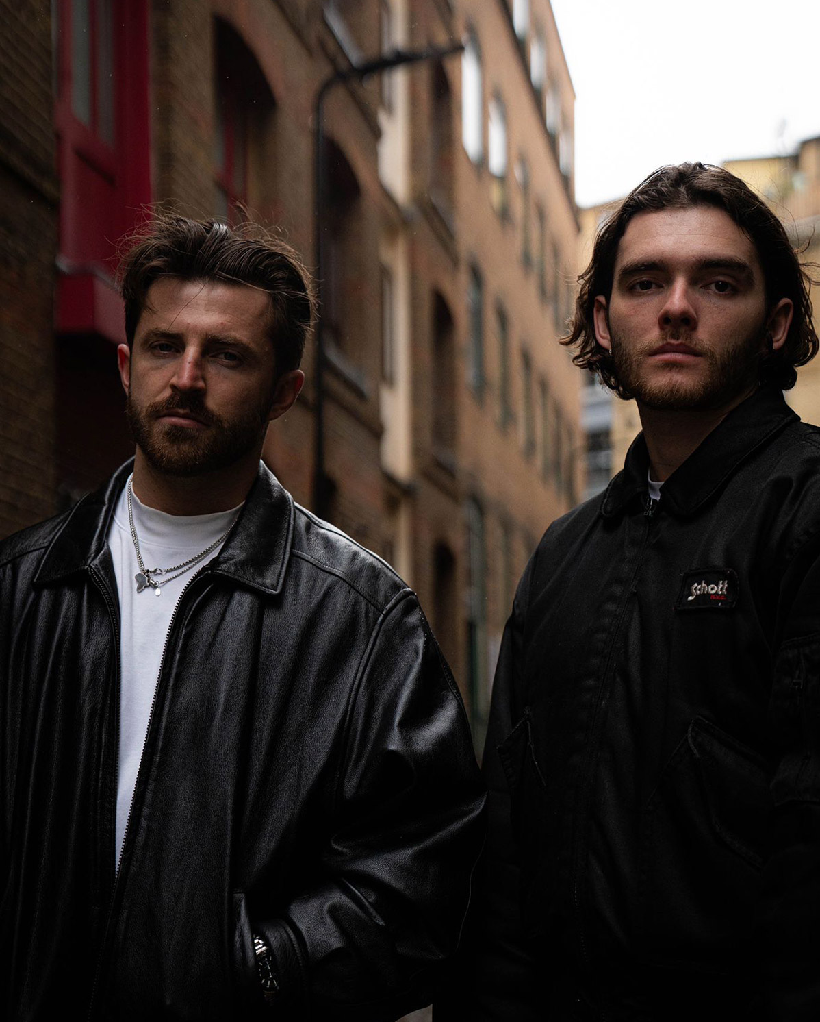 Two men wearing dark jackets stand closely together in a narrow alley with brick buildings on either side. The lighting is dim, and both have serious expressions.
