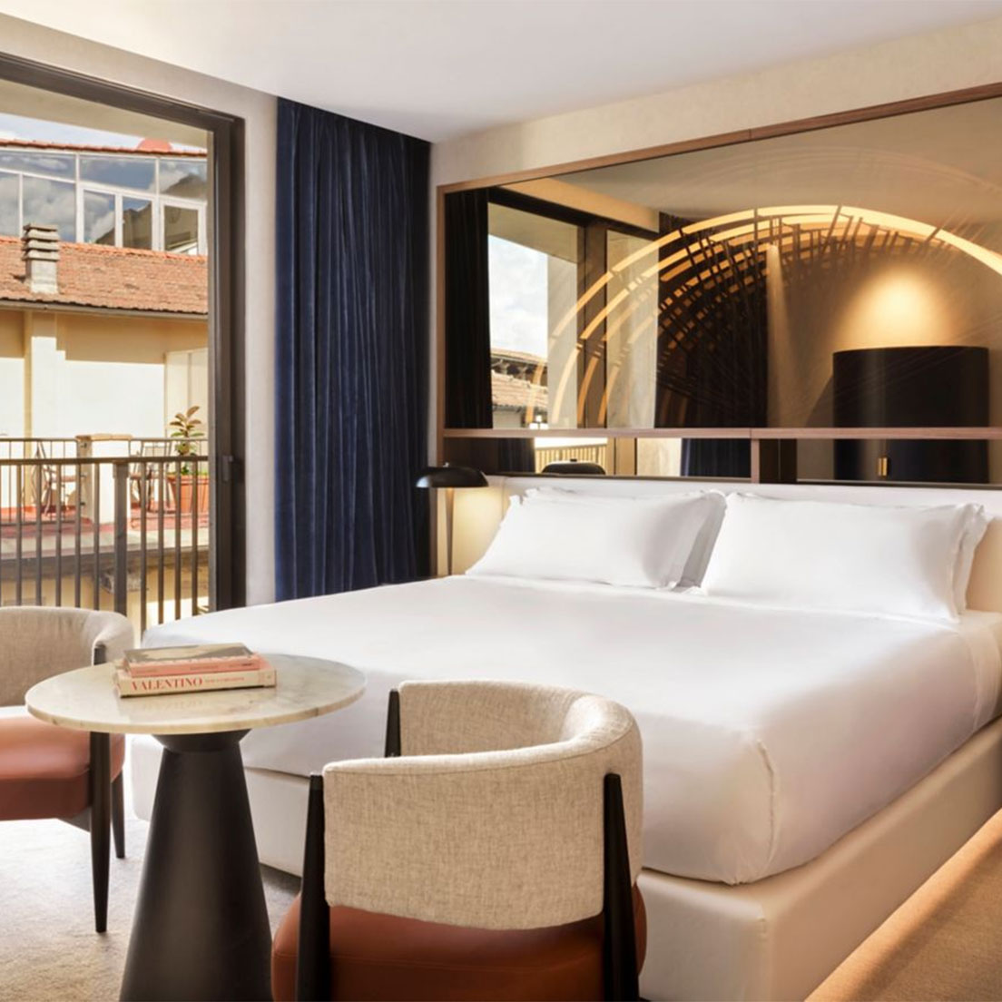 Modern hotel room with a large bed, two beige chairs, a round table with books, a floor lamp, and a large window opening to a balcony with building views. A decorative wall mirror is above the bed.