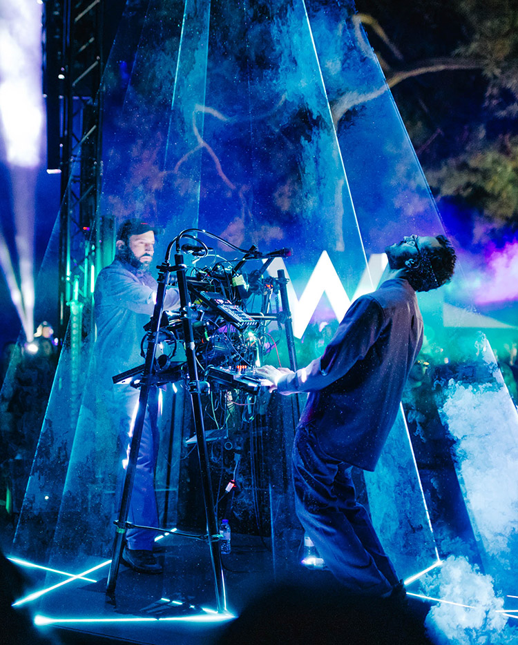 Two musicians perform on stage surrounded by blue lights and fog, standing inside a transparent triangular structure with electronic equipment between them; a crowd is visible in the background.