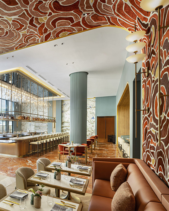 A stylish restaurant interior with modern decor, featuring a bar with high chairs, marble flooring, green columns, plush seating, and bold red and gold patterned walls and ceiling. Tables are set for dining.