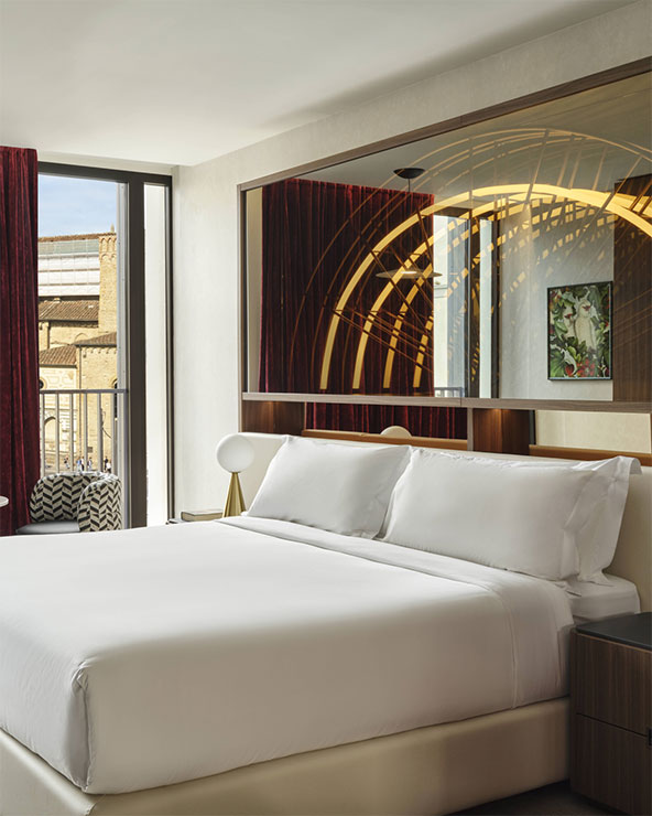 A modern hotel room with a neatly made bed, white linens, a large mirror above the headboard, contemporary lamps, red curtains, and a window with a view of a building outside.