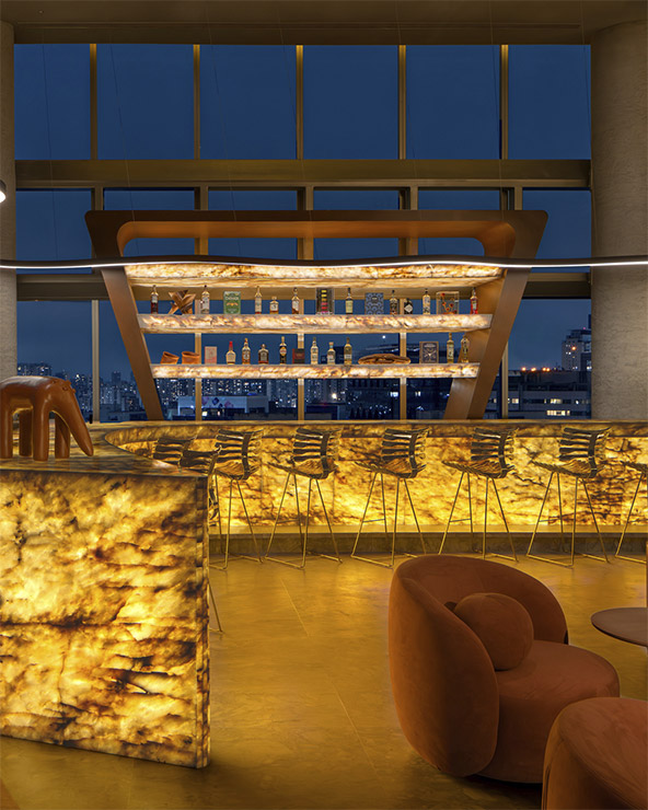 A stylish bar with glowing amber-colored backlit counters, modern barstools, curved orange lounge chairs, and large windows revealing a city skyline at night. Bottles are displayed on illuminated shelves behind the bar.