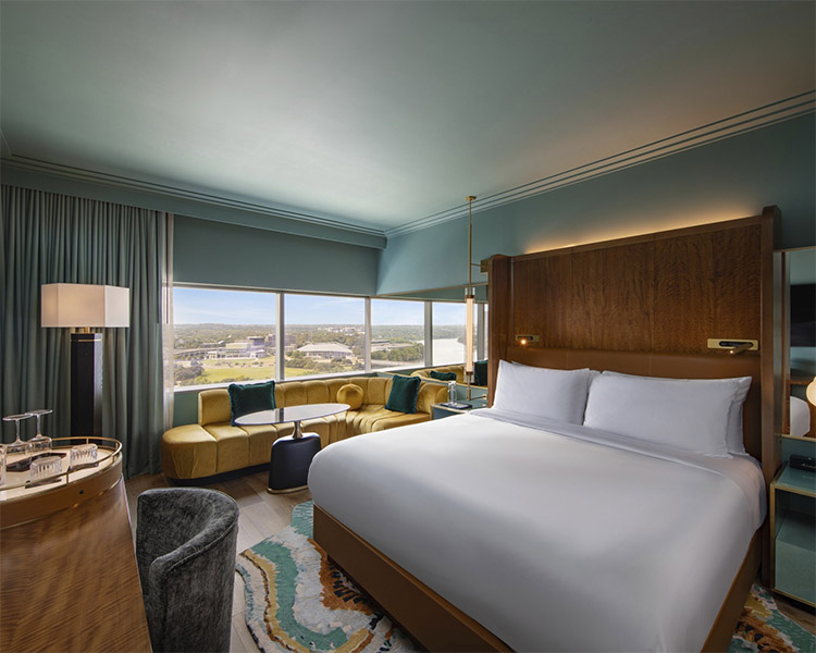 A stylish hotel room with a large bed, a curved yellow sofa by wide windows, a modern armchair, a lamp, and a scenic view of buildings and greenery outside. The room has soft blue and wood tones.