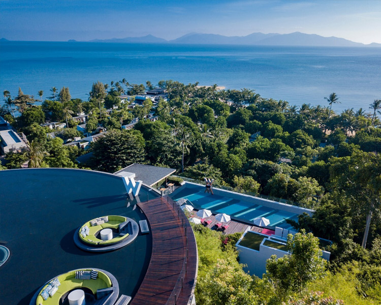 Aerial view of a luxury resort surrounded by lush greenery, with a rooftop lounge, swimming pool, and ocean in the background. Mountains are visible across the water under a clear blue sky.