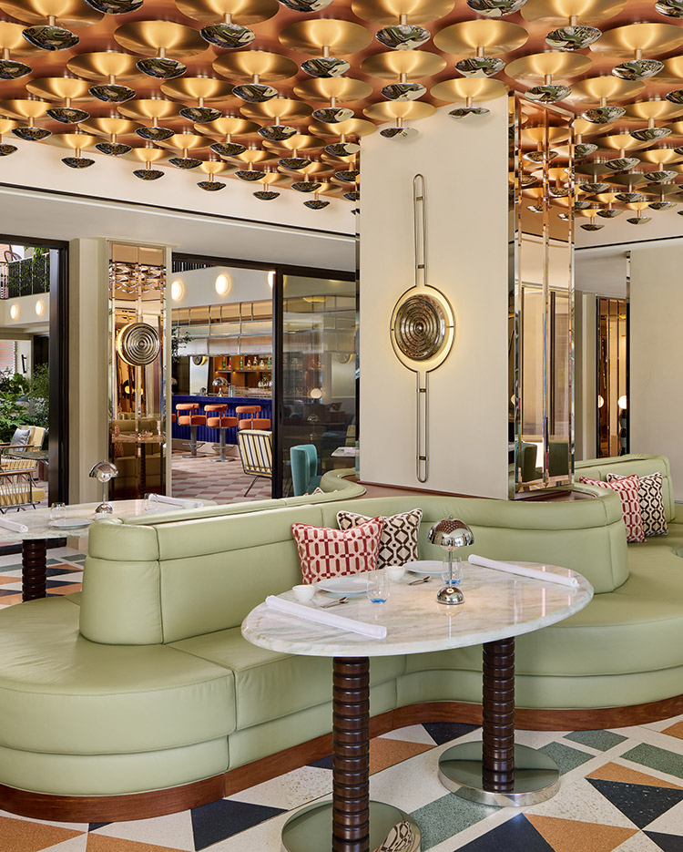 Chic restaurant interior with mint-green curved sofas, marble tables, geometric-patterned floor, gold decorative ceiling, and retro-style wall mirrors. The space feels modern and luxurious with unique lighting and accent pillows.
