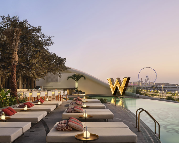 A modern rooftop pool area with lounge chairs, small tables with glowing lamps, and a large illuminated 