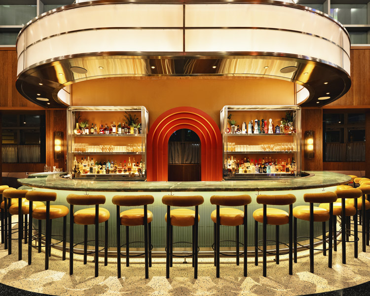 A stylish, retro-inspired bar with a curved counter lined with yellow cushioned stools. Shelves behind the bar display bottles and glassware, with a bold red arch feature in the center, warm lighting, and a mid-century modern design.