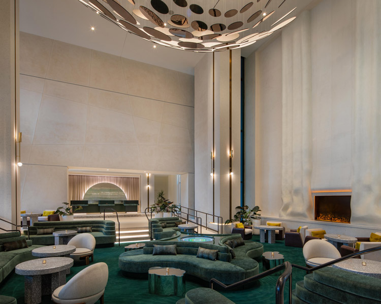 A modern, spacious hotel lobby with green velvet seating, round tables, a large abstract ceiling sculpture, tall white walls, and a contemporary fireplace. The lighting is soft and elegant.