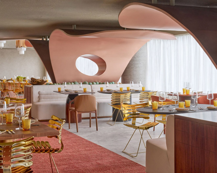 A modern restaurant interior with sculptural pink ceiling elements, gold and wood chairs, round tables set with yellow glasses, and a plush white booth by large windows with sheer curtains.
