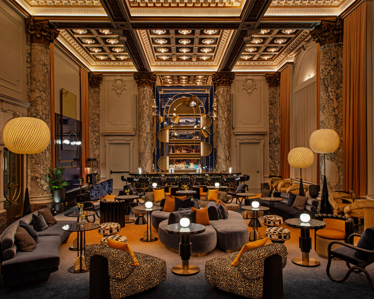 Luxurious hotel lounge with elegant marble columns, high ornate ceiling, warm lighting, plush sofas, patterned armchairs, round tables, and a central bar stocked with bottles, creating a sophisticated and inviting atmosphere.