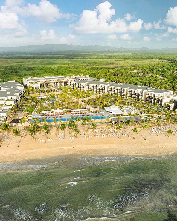 Aerial view of a large beachfront resort with multiple buildings, palm trees, pools, lounge chairs, and sandy beach, surrounded by lush greenery and mountains in the background.