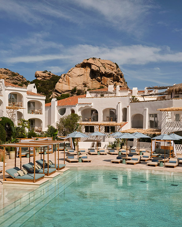 A luxurious resort with white Mediterranean-style buildings, sun loungers, and shaded cabanas surrounding a clear swimming pool, set against a backdrop of rocky hills under a blue sky.