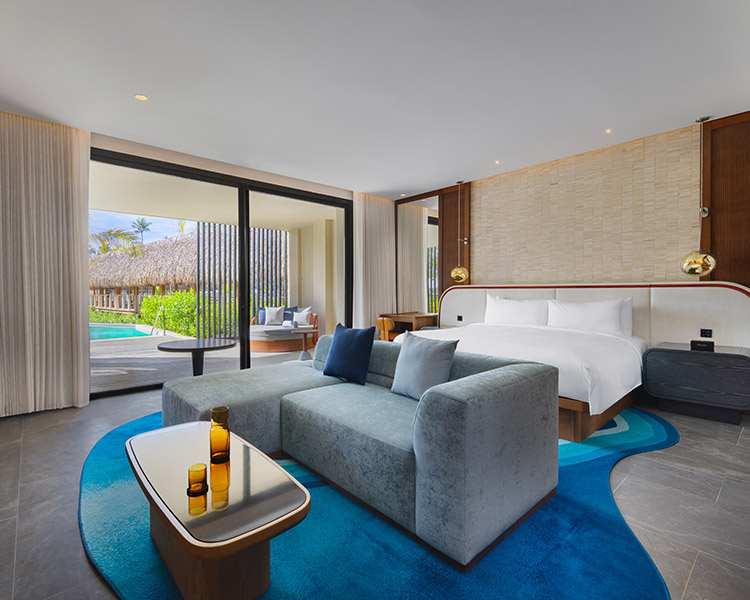 A modern hotel room with a large bed, light blue sofa, coffee table with amber glassware, and sliding glass doors opening to a private patio, green lawn, and pool outside. The decor is sleek with a blue patterned rug.