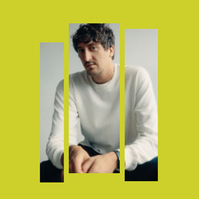 Un hombre con pelo ondulado y bigote, vestido con un jersey blanco, está sentado y mira a la cámara. La imagen está dividida en tres secciones verticales entre las que se ve un fondo amarillo brillante.