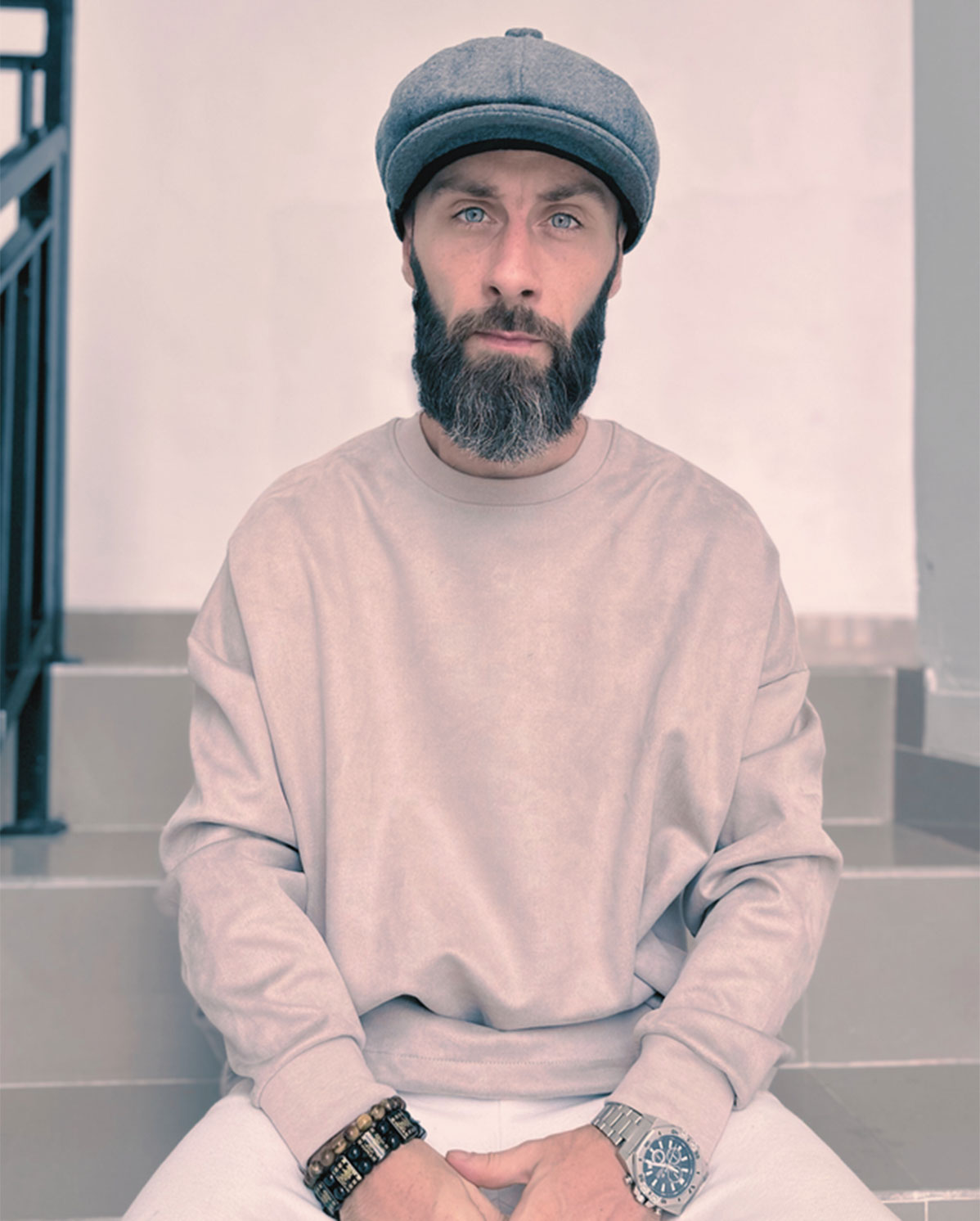 A bearded man wearing a grey flat cap, light pink sweatshirt, and several wrist accessories sits on stairs, looking directly at the camera with a neutral expression.